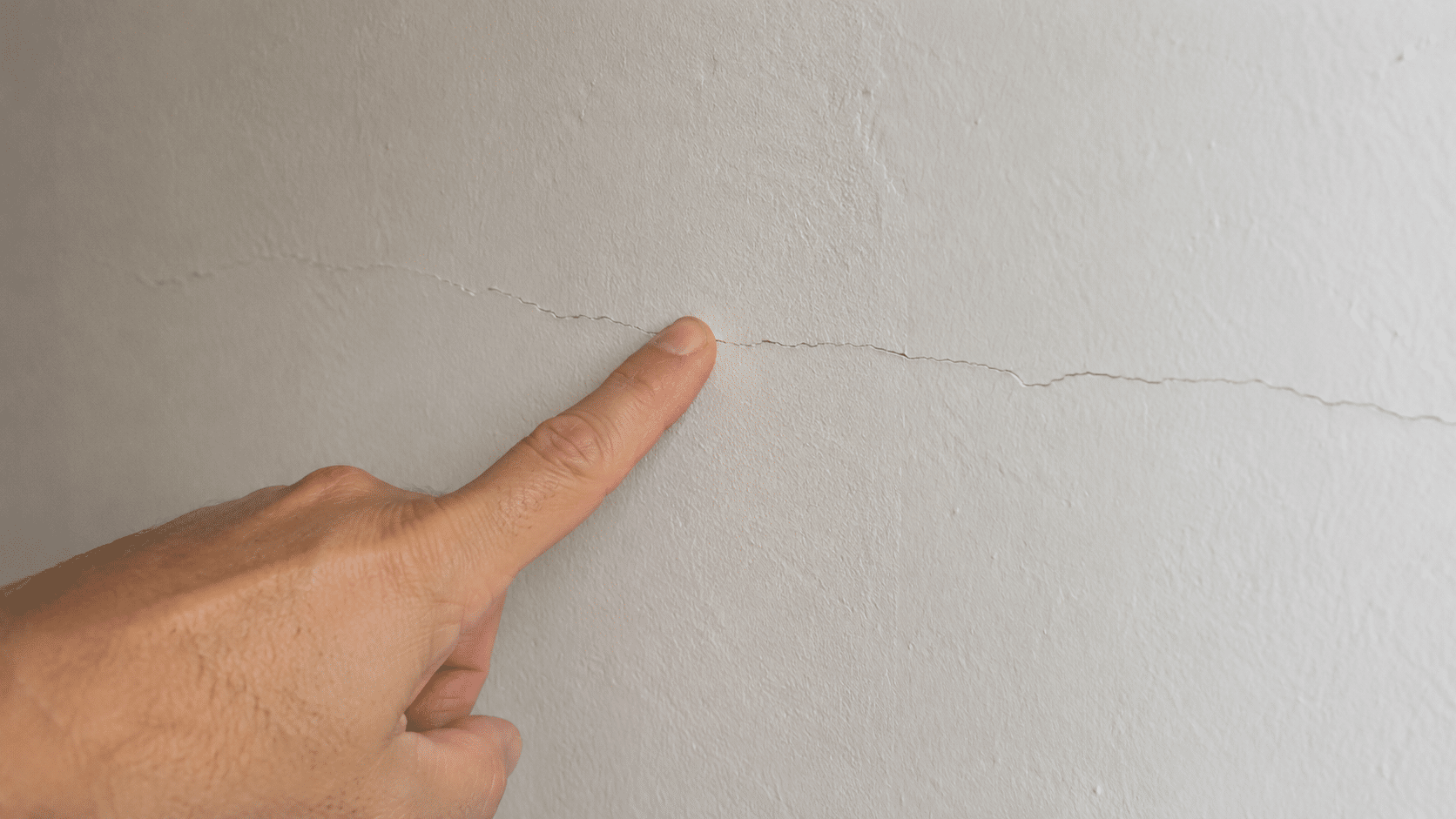 Main désignant une fissure discrète sur un mur, illustrant la frontière entre vice apparent et caché.