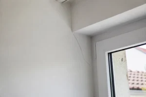 Fissure fine à l'angle d'une fenêtre sur un mur intérieur, désordre lié aux variations saisonnières du sol