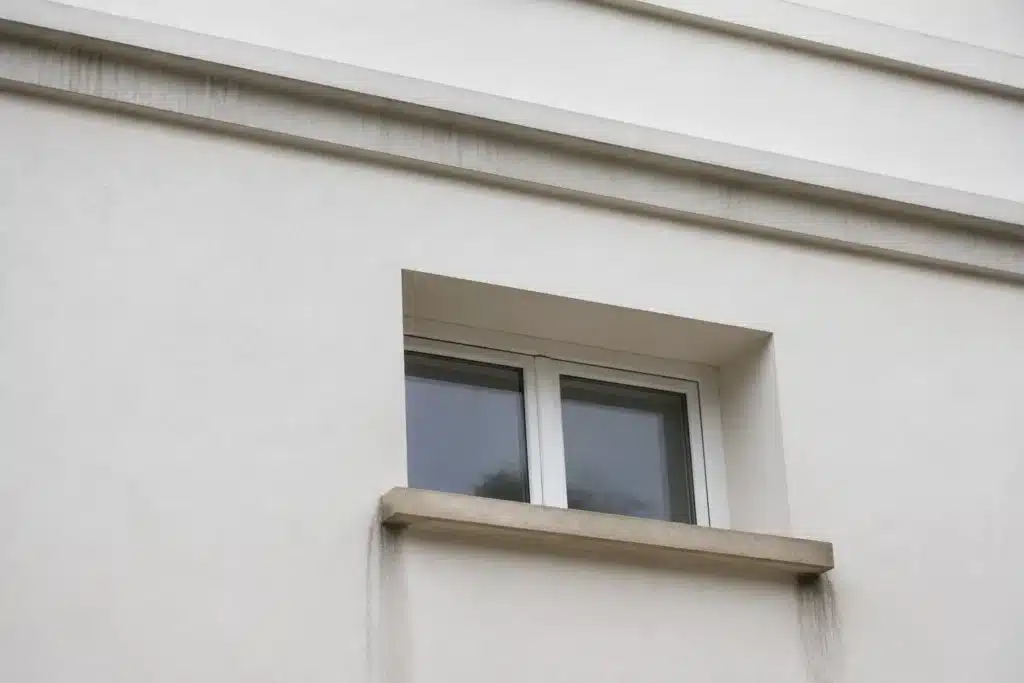 Appui de fenêtre sans goutte d'eau sous une façade fraîchement ravalée, avec coulures grises sur l'enduit clair sous les angles de l'appui
